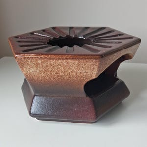 May include: A hexagonal ceramic stand featuring a dark brown glazed finish that transitions into a lighter tan speckled texture near the top. The top surface has a central circular opening surrounded by radiating grooves, and the base has side cutouts.