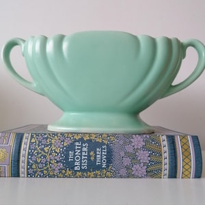 May include: A pale green ceramic vase with two handles and a fluted design. The vase is placed on a book titled "The Brontë Sisters Three Novels". The book has a floral pattern.
