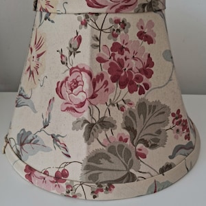 May include: A fabric lampshade with a floral design. The shade has a cream-coloured base, adorned with a pattern of pink and burgundy roses, leaves, and berries. It has a classic, tapered shape, suitable for a table lamp.