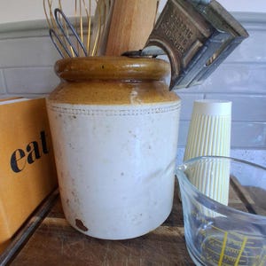 May include: A selection of kitchen utensils, including a whisk, wooden spoon, and a vintage jar. A measuring jug and a yellow ribbed container are also visible. The jar has a brown top and a white body.