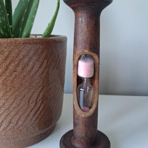 May include: A vertical hourglass featuring a dark brown wooden frame with a weathered texture and a metallic base and top. The central glass chamber contains fine pink sand, with a portion of the sand currently resting in the bottom bulb.