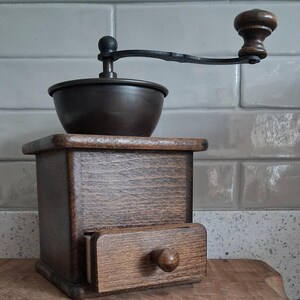 May include: A manual coffee grinder featuring a dark brown wooden box base with a small pull-out drawer and a round wooden knob. A dark metal bowl sits on top, equipped with a long black metal handle that has a small wooden knob at the end.