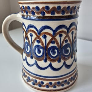 May include: A ceramic mug with a cream-coloured base featuring hand-painted blue and brown decorative patterns. The design includes horizontal bands of wavy lines, dots, and repeating arched motifs that wrap around the cylindrical body.