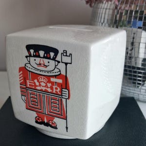 May include: A white ceramic cube-shaped money box featuring a crackle glaze finish. The front displays a graphic illustration of a British guard in a red uniform with the letters E and R flanking a crown symbol. The guard holds a tall staff.