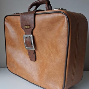 May include: A tan suitcase with a textured finish and dark brown piping. It features a dark brown handle and a central vertical strap with a silver-toned metal buckle. The word SPARTAN is printed in gold on an oval badge on the front strap.
