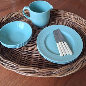 May include: A woven serving tray displays turquoise tableware: plates, bowls, and a jug. Three knives with cream handles are on a plate. The items are arranged on a wooden table.