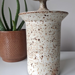 May include: A cylindrical ceramic canister with a speckled off-white glaze and dark brown spots. The canister features a wide, flared rim and a matching lid with a tall, mushroom-shaped handle centred on top, creating a distinct silhouette.