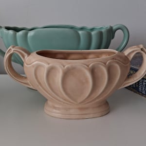 May include: Two ceramic planters. The front planter is a light peach colour with two handles and a scalloped design. The back planter is a teal colour with a scalloped rim and two handles. Both planters are on a white surface.