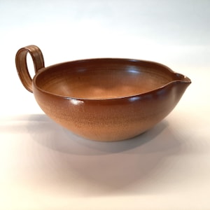 May include: A brown ceramic bowl with a single handle. The bowl has a slightly rounded shape and a smooth, glazed finish.