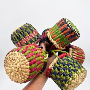 Op de afbeelding: Een verzameling geweven manden in verschillende kleuren, waaronder groen, roze, blauw en oranje. Elke mand heeft een cilindrische vorm met een geweven handvat. De manden zijn gemaakt van natuurlijke vezels en hebben een getextureerd uiterlijk.