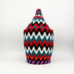 May include: A woven basket with a conical lid. The basket features a vibrant zigzag pattern in red, teal, black, white, and orange. The base is purple, and the top of the lid has a purple and white stripe.