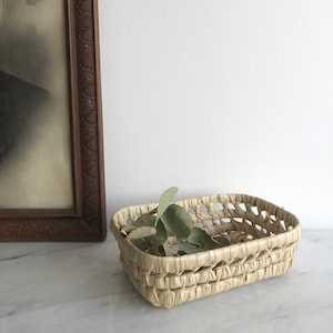 May include: A rectangular woven basket made of natural straw with a light brown color. The basket is empty and has a few green leaves inside.