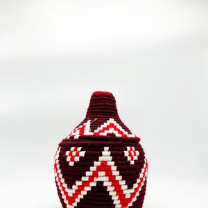 Puede incluir: Una cesta tejida de color granate con tapa. La cesta presenta un patrón geométrico de zigzags rojos y blancos. La tapa está rematada con un acento rojo. La cesta es probablemente hecha a mano y adecuada para guardar o decorar.