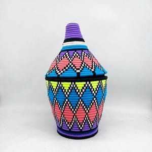 May include: A decorative woven basket with a lid. The basket features a vibrant geometric pattern in pink, blue, black, white, and yellow. The lid is cone-shaped and purple. The basket is a unique storage solution.