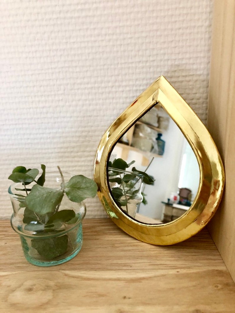 May include: A gold teardrop-shaped mirror with a beveled edge. The mirror is reflecting a room with a plant in a glass vase.
