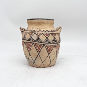 May include: A beige ceramic vessel with two small handles, featuring a hand-painted geometric pattern of black and reddish-brown triangles and diamonds. The pot has a wide mouth with a dark rim and a textured, matte finish on its exterior surface.