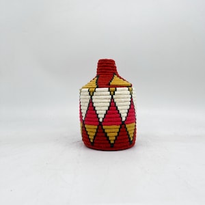 May include: A handwoven storage basket with a lid. The basket features a geometric diamond pattern in red, white, and gold. The lid and top section are primarily red. The basket is made of natural fibers.