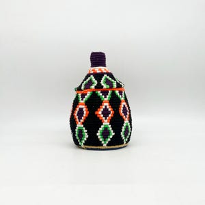 May include: A hand-woven storage jar with a lid. The jar is black with a repeating pattern of orange, green, and purple diamonds. The lid and the top of the jar are purple. The jar is sitting on a white surface.