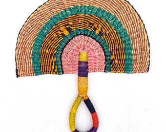 May include: A woven fan with a colorful, striped design. The fan is made of natural materials and has a pink, orange, green, and blue color scheme. The fan has a woven handle.