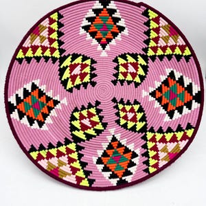 Op de afbeelding: Een ronde, geweven mand met een roze midden en een donker bordeauxrode rand. De mand heeft geometrische patronen in zwart, wit, geel, oranje en groen. Het ontwerp is symmetrisch, met herhalende vormen die vanuit het midden uitstralen.