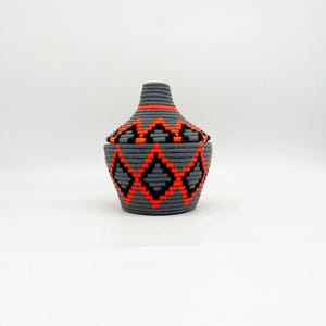 May include: A gray woven basket with a lid, featuring a geometric pattern in black and orange. The basket has a cylindrical shape with a slightly tapered top. The lid mirrors the pattern on the basket's body.
