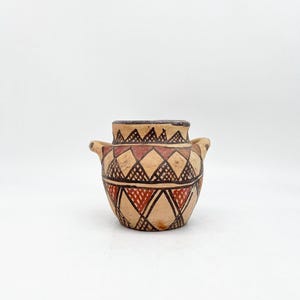 Puede incluir: Una pequeña olla de barro marrón con un patrón geométrico de triángulos rojos, negros y blancos. La olla tiene dos pequeñas asas a los lados.
