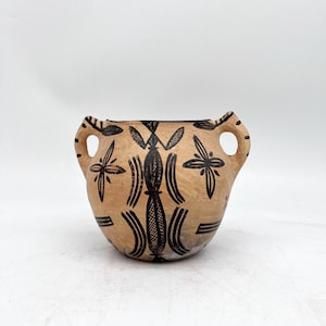 May include: A tan ceramic vessel with two side handles, featuring hand-painted black geometric patterns including cross-hatched vertical shapes, star-like motifs, and horizontal lines encircling the rounded body of the pottery piece.