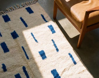Puede incluir: Una alfombra rectangular de color crema con un patrón geométrico de rectángulos y líneas azules. La alfombra tiene una textura peluda y una franja en un extremo. Un sillón de cuero marrón con brazos de madera está en el fondo.