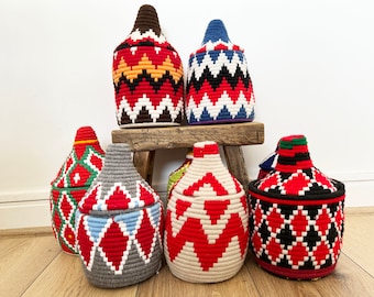 Könnte beinhalten: Sechs bunte gewebte Aufbewahrungskörbe mit Deckeln, die geometrische Muster in Rot, Weiß, Schwarz, Blau, Grün und Braun aufweisen. Die Körbe sind auf einem Holzhocker und einem Holzboden angeordnet.