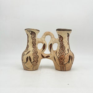 May include: A white ceramic vase with two openings, decorated with brown and red painted designs. The vase has a unique shape with a central opening and two smaller openings on either side.