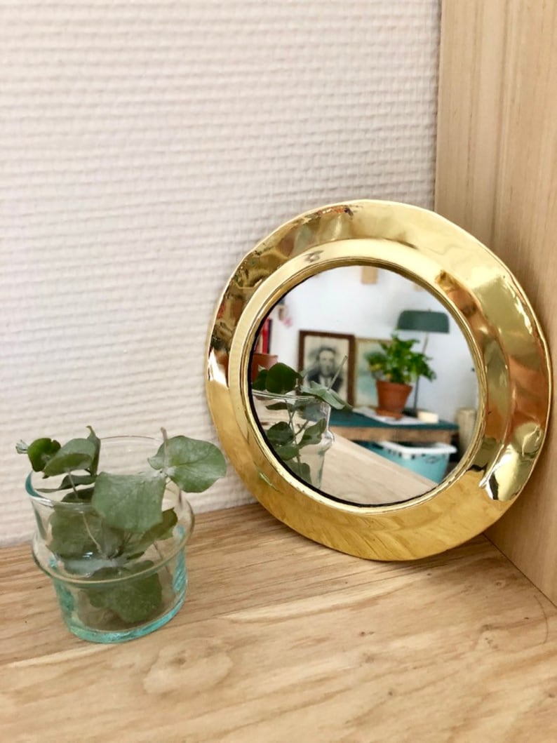 Puede incluir: Un peque&ntilde;o espejo redondo de lat&oacute;n con un marco ancho y redondeado. El espejo refleja una habitaci&oacute;n con plantas y muebles.