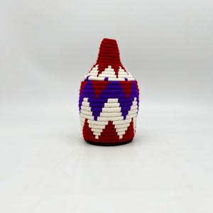 May include: A woven basket with a lid, featuring a geometric pattern. The basket's design incorporates red, purple, and white colors. The basket is cylindrical with a conical lid.