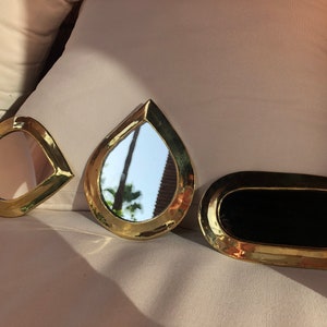May include: Three gold-toned decorative mirrors with different shapes. The mirrors are on a white surface. The largest mirror is oval-shaped and has a black reflective surface. The other two mirrors are teardrop-shaped and have a silver reflective surface.