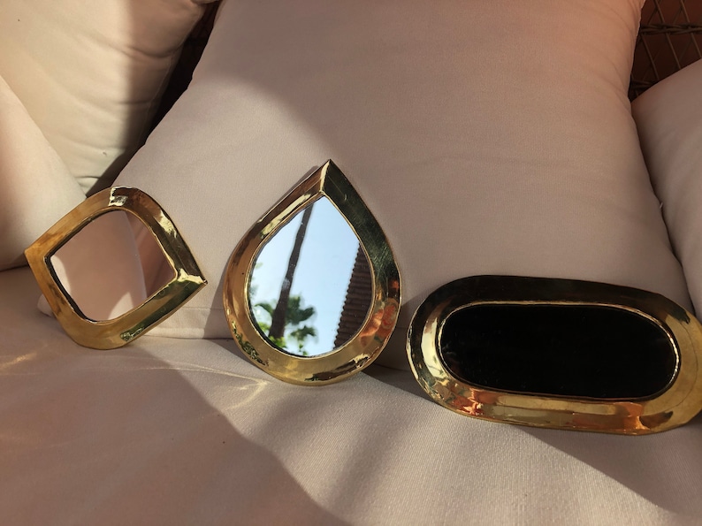 May include: Three gold-toned decorative mirrors with different shapes. The mirrors are on a white surface. The largest mirror is oval-shaped and has a black reflective surface. The other two mirrors are teardrop-shaped and have a silver reflective surface.