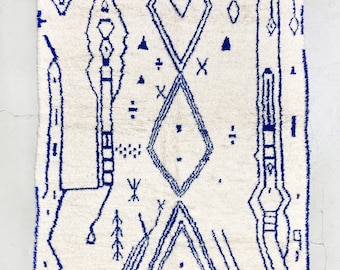 May include: A white rug with a blue geometric pattern. The pattern features diamonds, lines, and other abstract shapes. The rug has a fringe border.