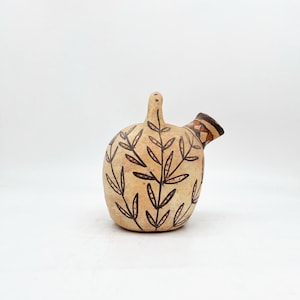 May include: A small, beige ceramic pitcher with a brown leafy design. The pitcher has a spout and a handle.