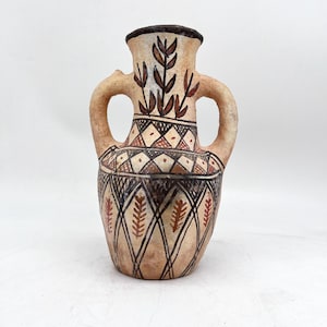 May include: A tan ceramic amphora vase featuring two side handles and a narrow neck. The exterior is decorated with hand-painted black and reddish-brown geometric patterns, including intersecting lines, diamonds, and stylized leaf motifs.