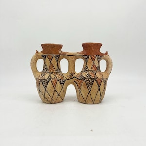 May include: A double-chambered ceramic vessel featuring two connected spouts and handles. The beige clay body is decorated with a hand-painted geometric diamond pattern in black and reddish-brown, with a star-like motif between the two chambers.