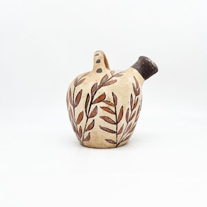May include: A beige ceramic pitcher with a handle and a spout. The pitcher is decorated with brown leaf patterns and small black dots. The spout and handle are brown.