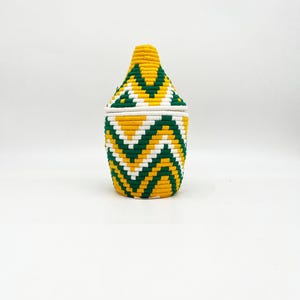 以下が含まれることがあります： 蓋付きの織り収納バスケット。バスケットは円筒形で、円錐形の蓋が付いています。 黄色、緑、白のシェブロン模様が繰り返されています。 背景は無地の白です。