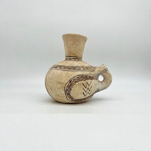 May include: A small, cream-colored ceramic jug with a brown handle and geometric designs. The jug has a narrow neck and a rounded body.