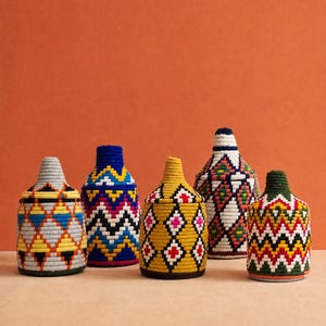 Peut inclure: Cinq récipients cylindriques avec des couvercles coniques, chacun enveloppé de fil multicolore vibrant. Les motifs présentent des formes géométriques dans des tons de jaune, bleu, rouge, blanc, noir et vert, créant un aspect tissé texturé.