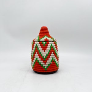 以下が含まれることがあります： 蓋付きの織りバスケット。バスケットは円筒形で、丸い底と円錐形の蓋が付いています。赤、白、緑のジグザグ模様が繰り返されています。バスケットは、しっかりと織られた素材で作られています。