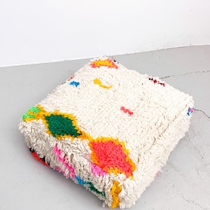 May include: A white fluffy square floor cushion with colorful geometric patterns in pink, yellow, green, blue, and red.