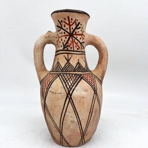 May include: A beige ceramic vase with two handles and a dark brown rim. The vase features geometric patterns in black and red, including a snowflake-like design near the top and diamond shapes. The vase has a textured, aged appearance.
