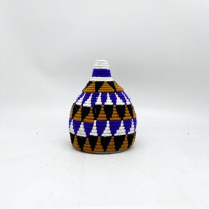 Puede incluir: Una pequeña cesta tejida con un patrón geométrico en tonos marrones, morados, blancos y negros. La cesta tiene forma de jarrón y tiene un fondo redondeado.