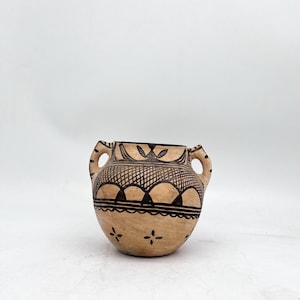 Könnte beinhalten: Eine handgefertigte, beige Keramikvase mit zwei Henkeln. Sie ist mit schwarzen geometrischen Mustern verziert, darunter Bögen und florale Motive am Rand und am Körper. Die Vase hat einen runden Boden und eine leicht ausgestellte Öffnung.