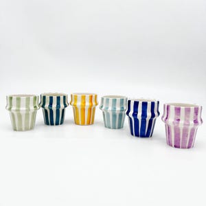 May include: Six small ceramic pots with vertical stripes in various colours. The pots are arranged in a row and feature stripes in green and white, navy and white, yellow and white, light blue and white, dark blue and white, and purple and white. The pots have a flared shape.