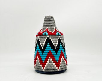 Könnte beinhalten: Ein grauer gewebter Aufbewahrungskorb mit einem geometrischen Muster in Rot, Blau, Schwarz und Weiß. Der Korb hat einen Deckel und einen runden Boden.