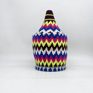 May include: A colorful woven basket with a geometric pattern in shades of pink, blue, white, yellow, and purple. The basket has a wide base and a narrow top, resembling a traditional Moroccan pot.